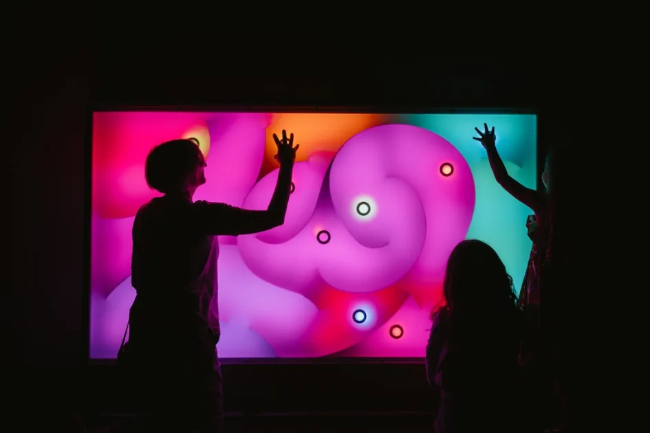 Silhouettes interact with a vibrant, glowing interactive screen in darkness.